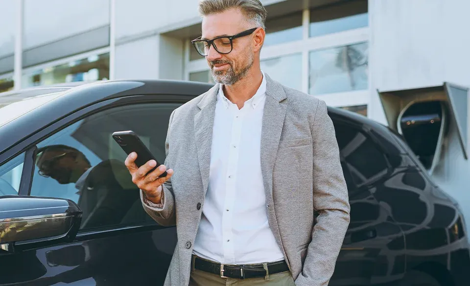 Mann im Anzug schaut auf Handy und vergleicht Fahrzeugangebote exklusiv f&uuml;r Firmen bzw. Gro&szlig;kunden. Im Hintergrund ist sein aktuelles neues E-Fahrzeug zu sehen. 